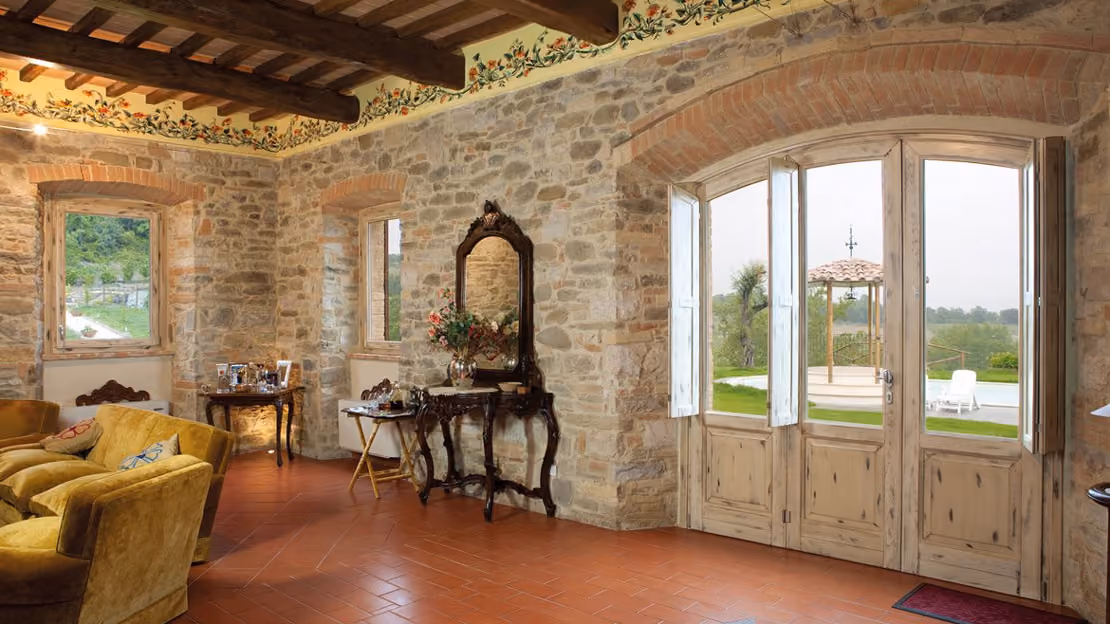 Cozy room with rustic stone walls, wooden ceiling beams, and terracotta floor tiles, featuring a large wooden arched door opening to a garden with a gazebo and pool, an ornate dark wood mirror with a flower vase on a small table, vintage furniture including mustard yellow armchairs, and a side table with various items near two windows.
