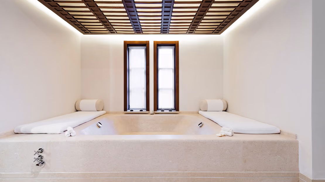 Luxurious beige stone bathtub with white cushioned padded headrests and rolled towels on both sides, placed under a decorative wood lattice ceiling and between two tall narrow windows with white blinds.