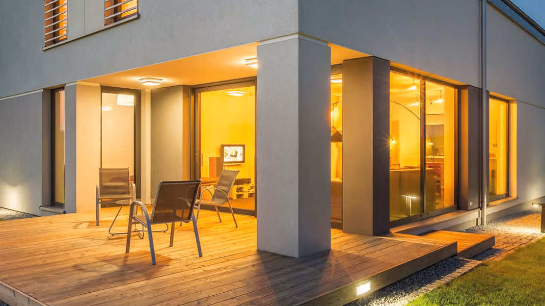 Modern house corner with large floor-to-ceiling windows and a wooden deck with three metal-framed chairs, illuminated warmly from inside during evening.