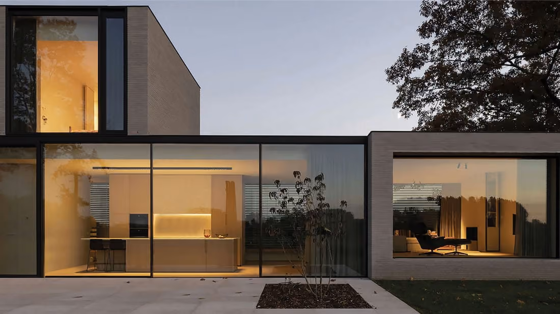 Modern house exterior at dusk showing large floor-to-ceiling glass windows revealing a warmly lit kitchen with bar stools on the left and a living room with a lounge chair on the right, surrounded by a concrete terrace and a small planter with a leafless shrub.