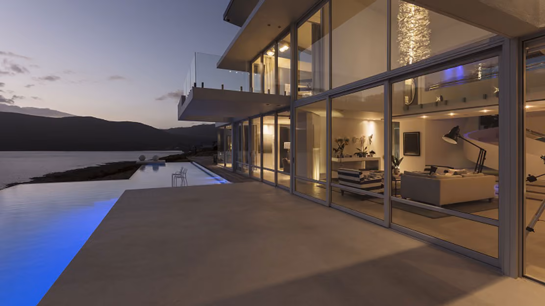 Modern house with large glass windows overlooking an infinity pool lit with blue lights, set against a backdrop of mountains and a body of water at dusk.