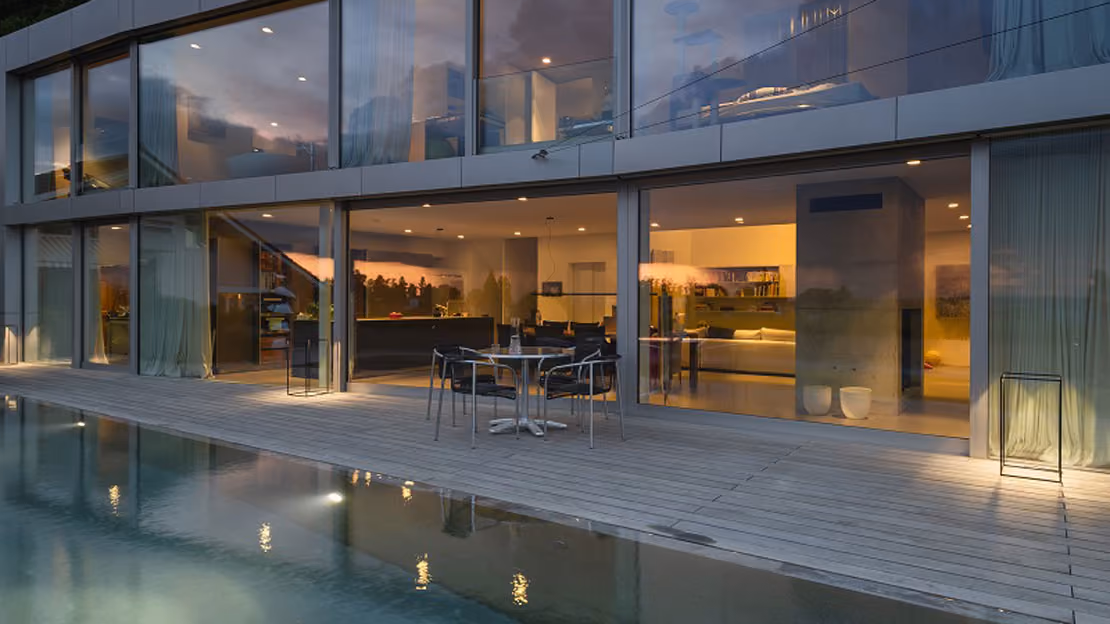 Modern house exterior at dusk with large floor-to-ceiling glass windows and sliding doors opening to a wooden deck with a small table and chairs beside a lit swimming pool.