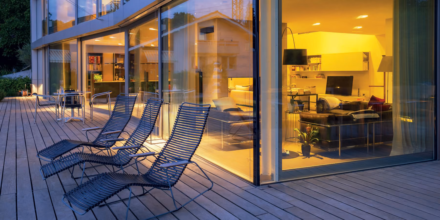 Modern home exterior at dusk with a wooden deck featuring three metal lounge chairs and a small round table with two chairs, large glass windows showcasing a warmly lit interior living room with a black leather sofa, bookshelves, and floor lamps.