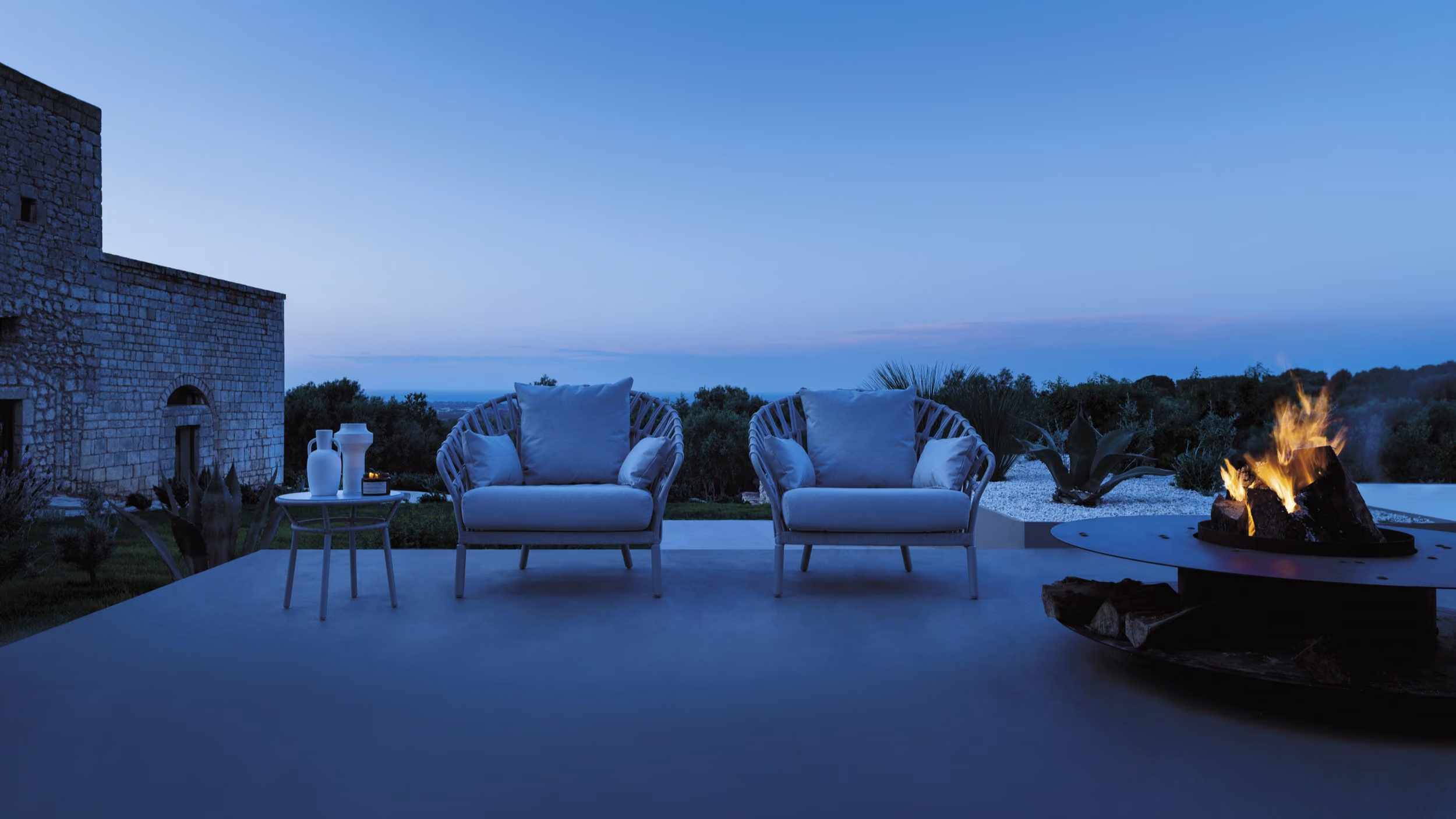 Outdoor seating area at dusk with two cushioned armchairs and a small table holding decorative vases, next to a circular fire pit with burning logs, set against a backdrop of greenery and a stone building.