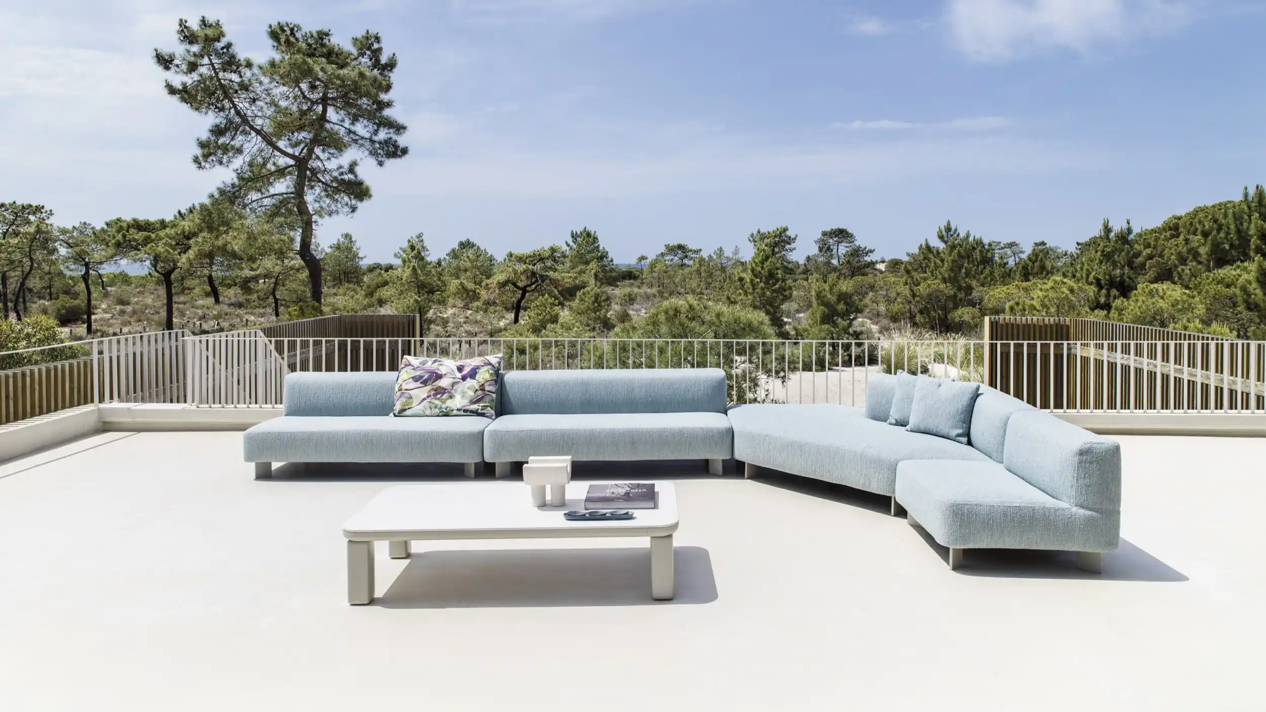 Modern outdoor terrace with a light blue L-shaped sectional sofa with cushions and a low white coffee table holding a book and decorative items, surrounded by greenery and trees under a clear sky.
