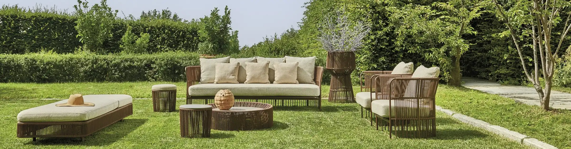 Outdoor patio furniture set on green grass, including a cushioned bench with a straw hat, a sofa with multiple beige cushions, two armchairs with beige cushions, two small round side tables, a round coffee table with a decorative vase, and a tall plant stand with flowers, surrounded by lush green bushes and trees.