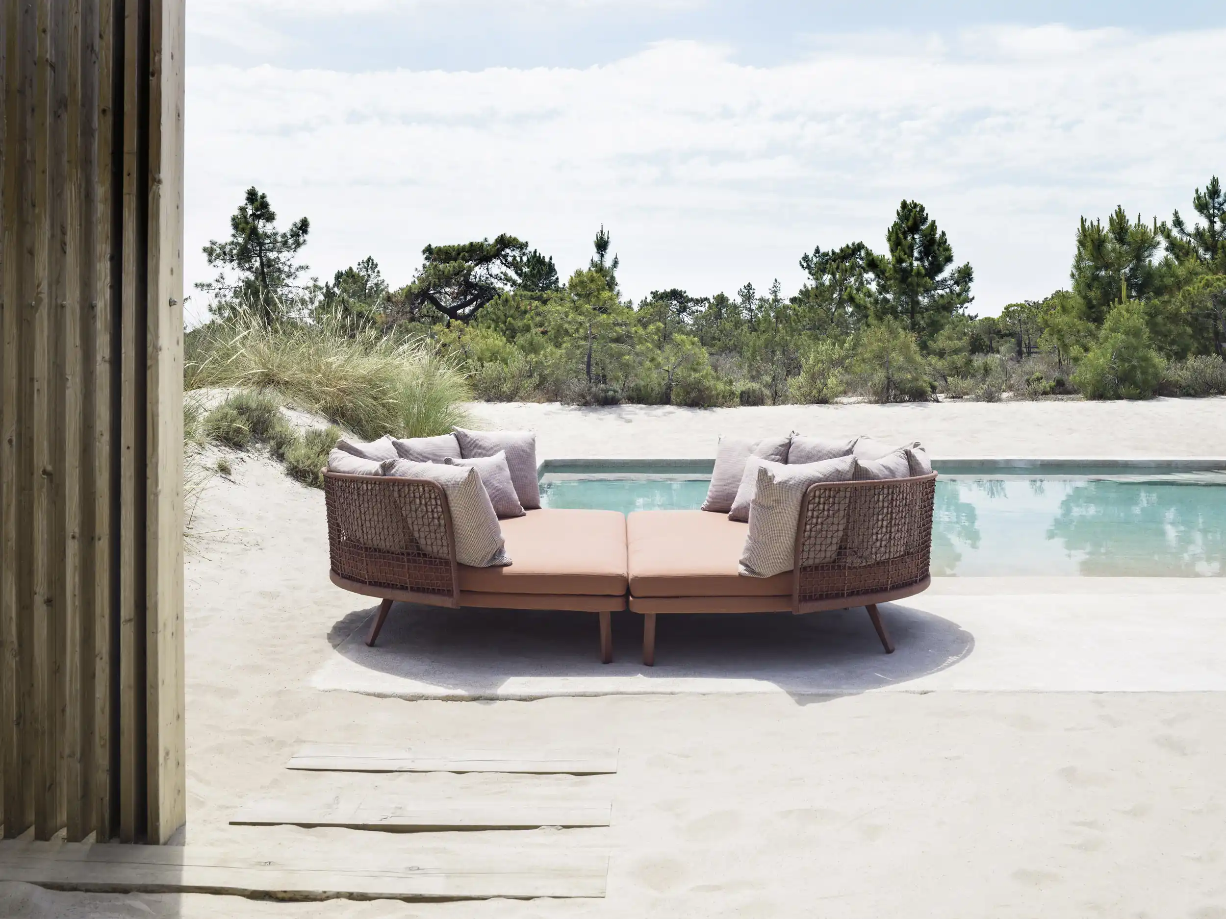 Outdoor seating area with two brown cushioned lounge chairs joined in a circular shape, placed on sand near a rectangular swimming pool surrounded by natural greenery under a cloudy sky.