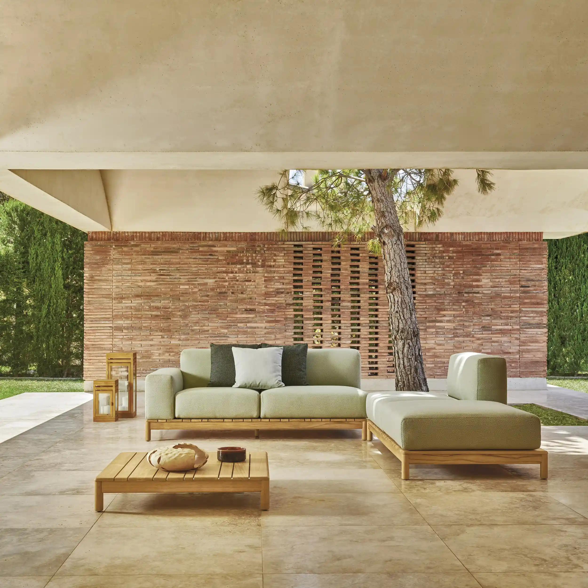 Modern outdoor patio with a light green cushioned wooden sofa set, including a sectional and a matching low coffee table holding decorative items, under a covered area with beige tile flooring, a red brick wall, and a tree growing through the roof structure.