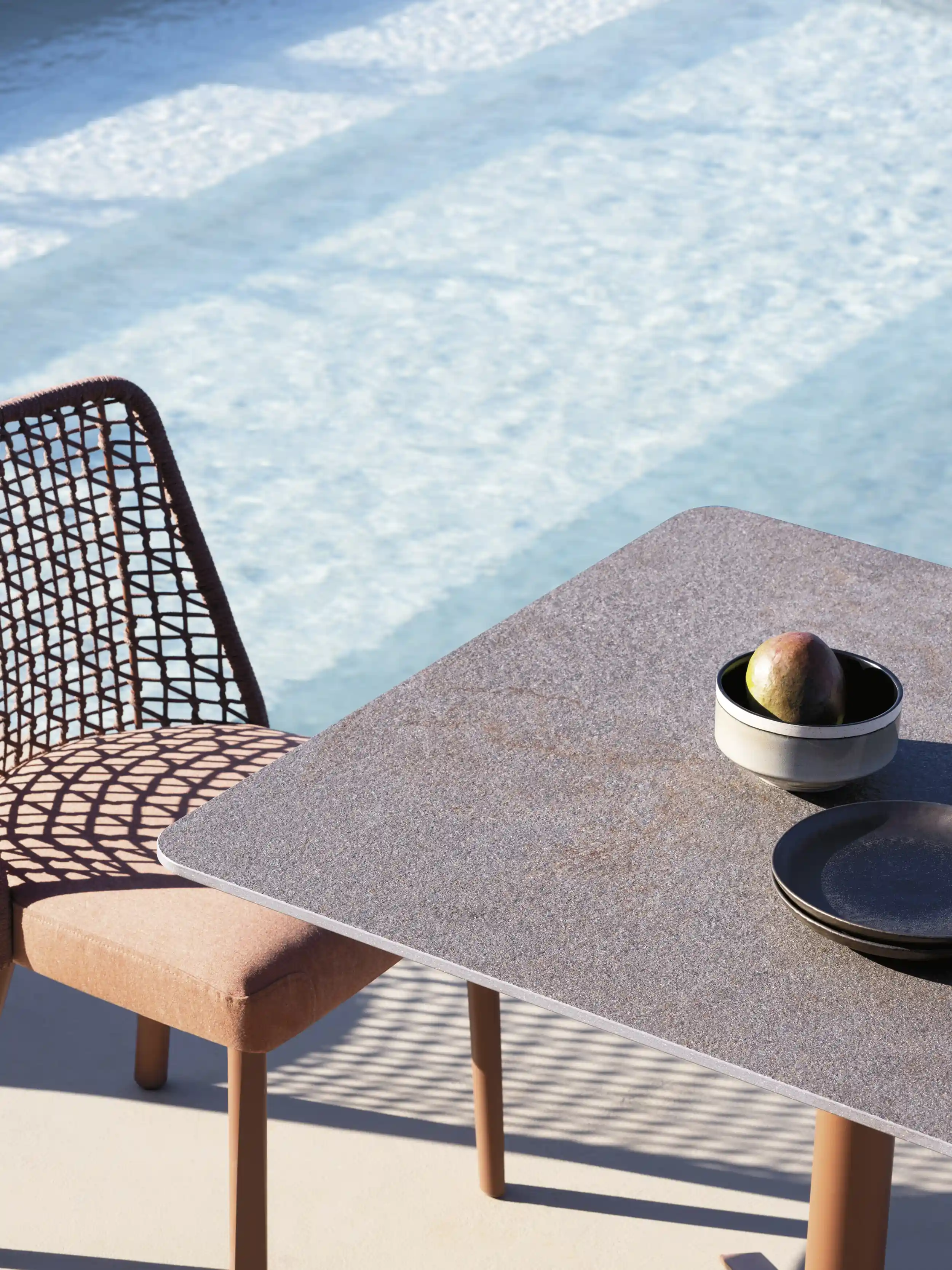 Outdoor setting with a beige cushioned chair with a woven back casting shadows on the ground, a gray stone table holding a bowl with a mango and two stacked black plates, with a light blue pool in the background.