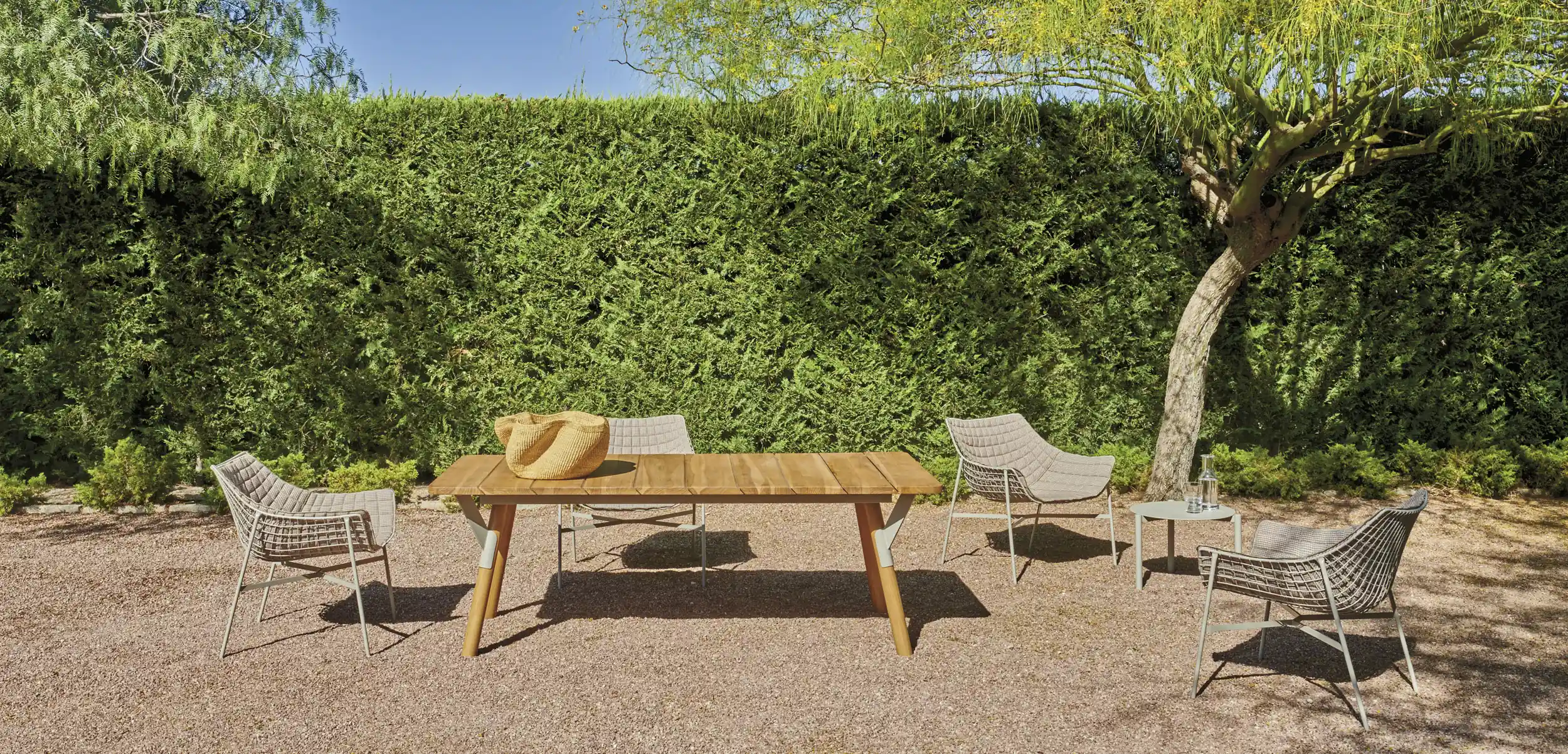 Outdoor seating area on gravel with a wooden table holding a woven basket, four cushioned metal chairs arranged around it, a small side table with a glass pitcher and cup, set against a tall green hedge and a tree on the right under a clear blue sky.