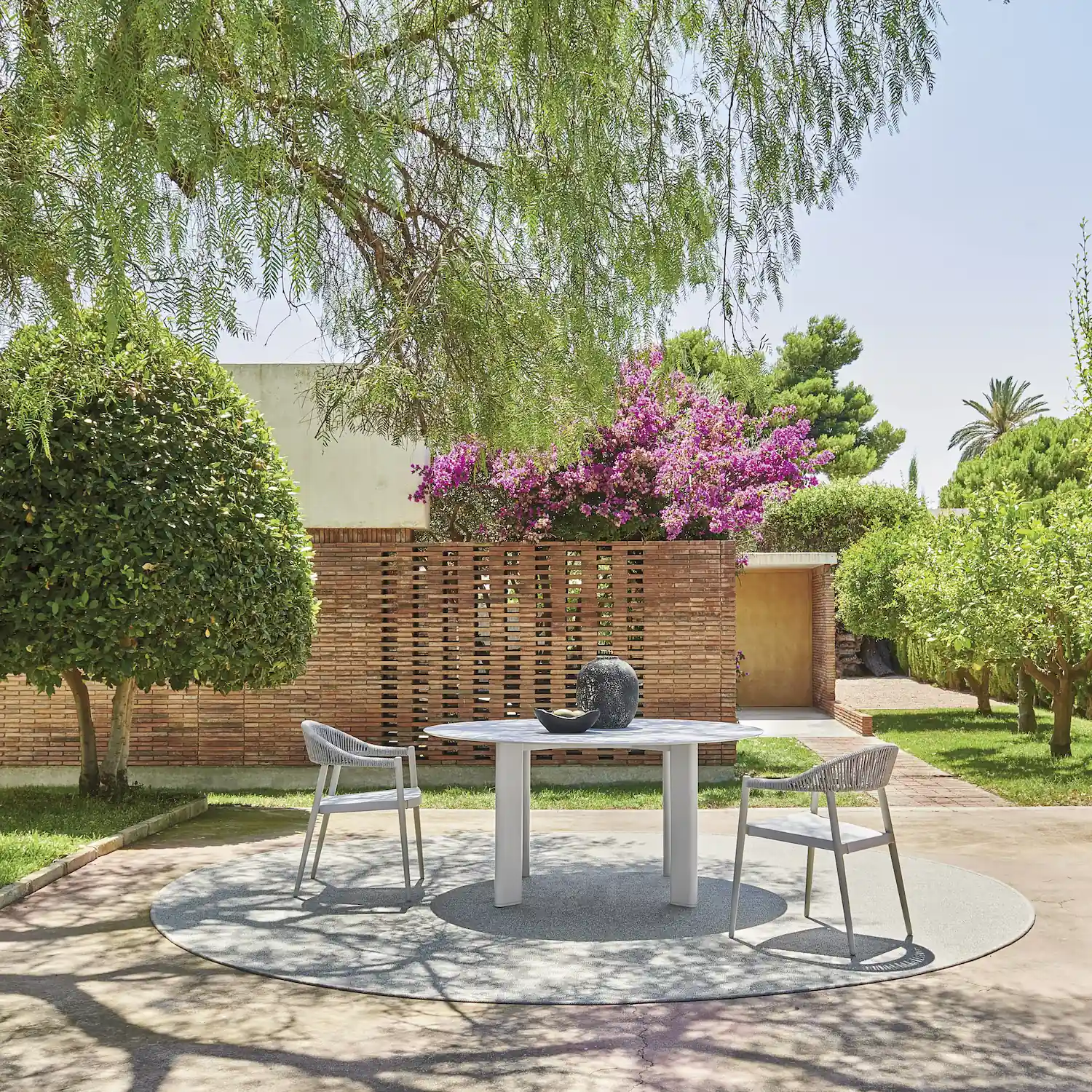 Outdoor patio area with a round white table and two gray woven chairs on a circular rug, surrounded by lush greenery, a brick wall, and flowering purple bougainvillea under a clear sky.