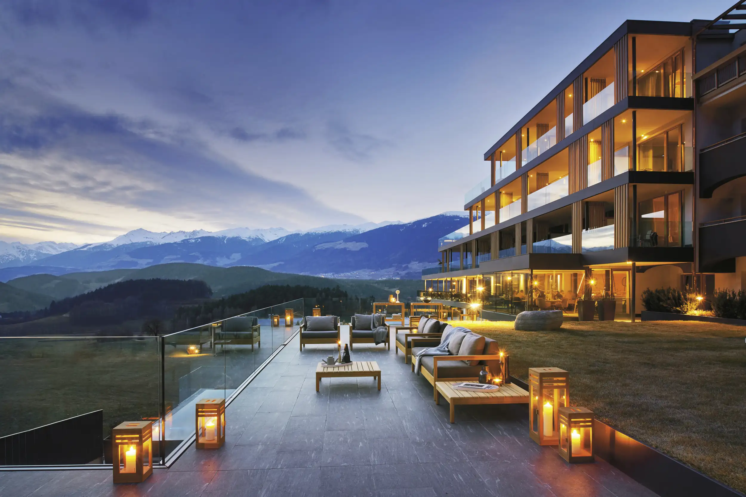 Modern luxury hotel terrace at dusk with lounge seating, wooden tables, lit lanterns, overlooking grassy area and mountains with snow-capped peaks in the background.