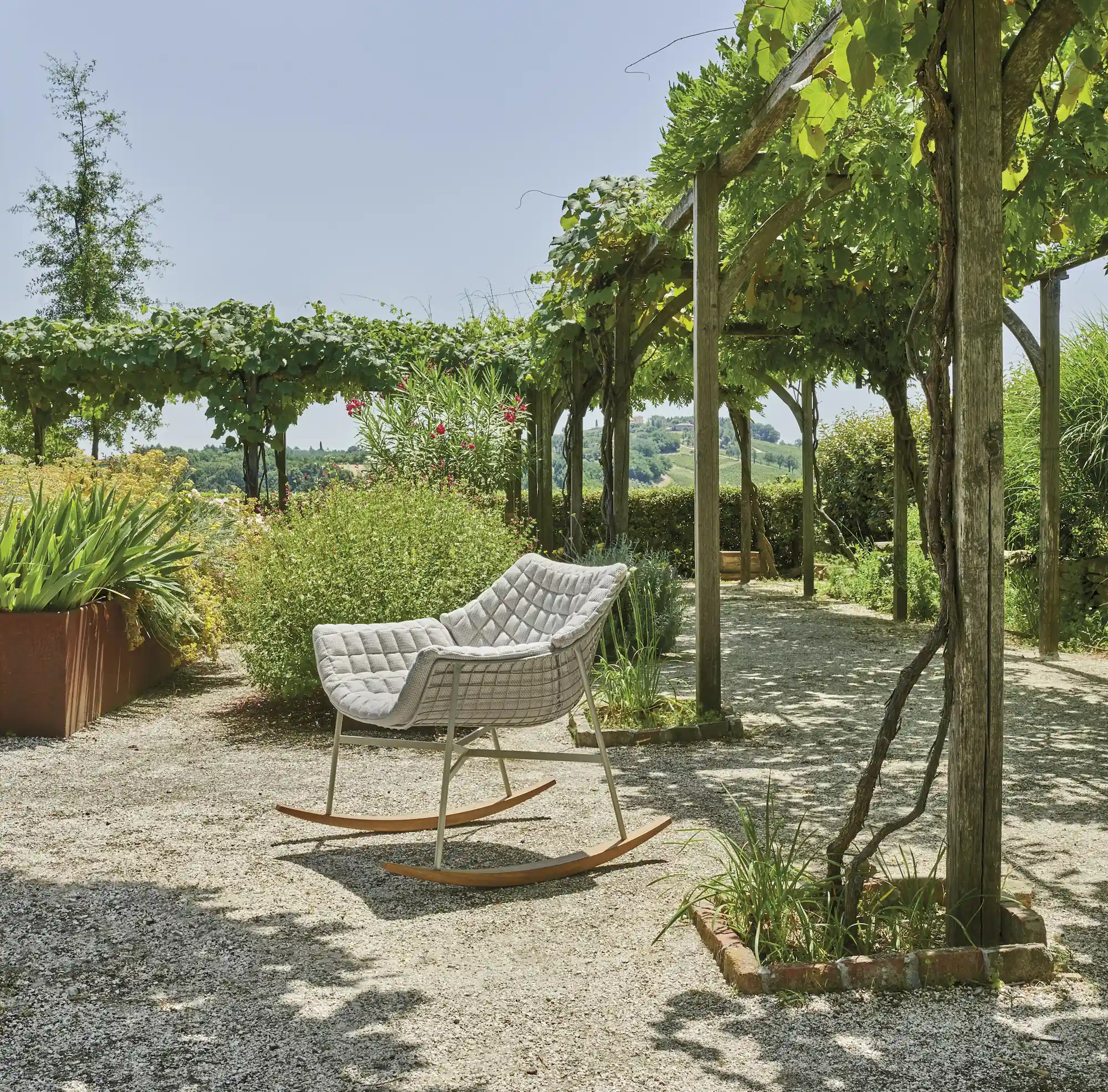 Modern cushioned rocking chair with a metal frame positioned on a gravel garden path under a wooden pergola covered in green vines, surrounded by lush plants and a scenic landscape in the background.