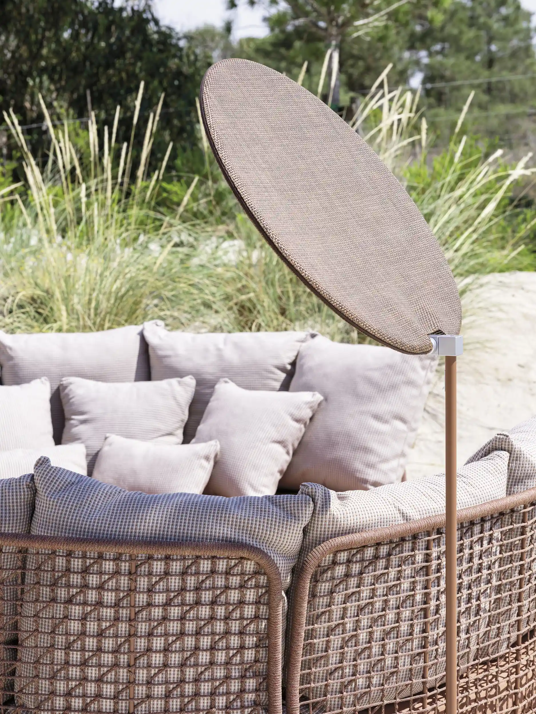 Curved outdoor woven rattan sofa with multiple light beige cushions and a tall, oval-shaped beige fabric shade positioned on a thin stand beside it, surrounded by greenery and grasses.
