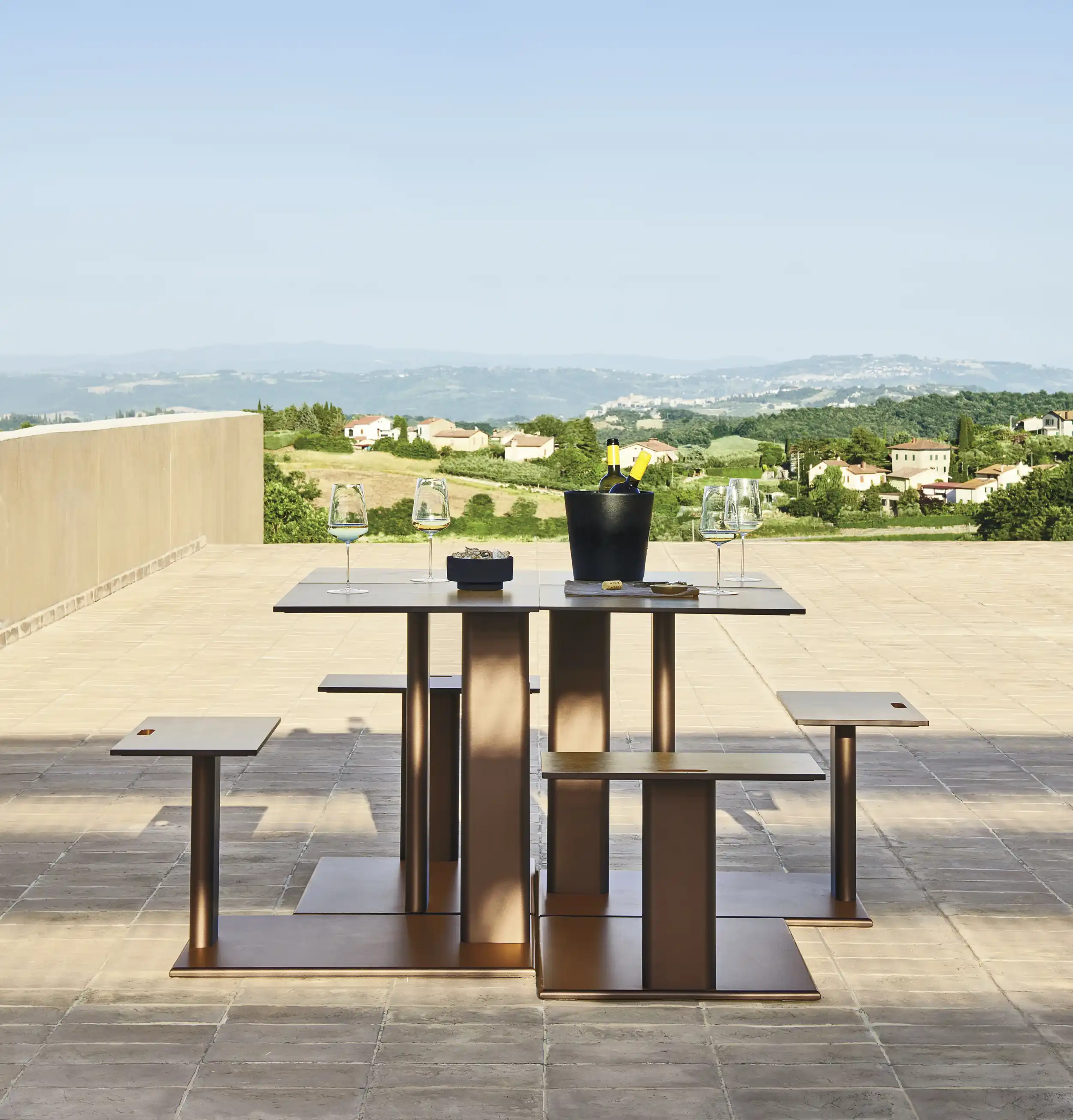 Modern bronze outdoor table with attached square stools set on a tiled terrace with wine glasses, a bucket with wine bottles, and a green countryside with houses in the background.