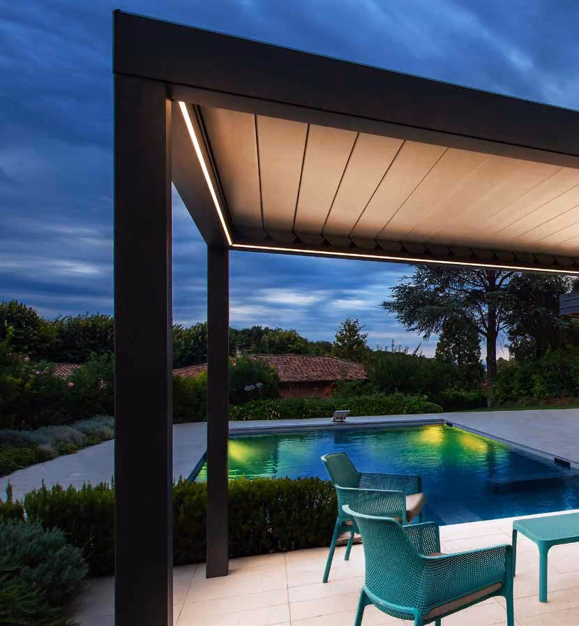 Modern outdoor patio with two teal chairs and a small table under a sleek pergola with recessed lighting, next to a lit swimming pool surrounded by greenery at dusk.