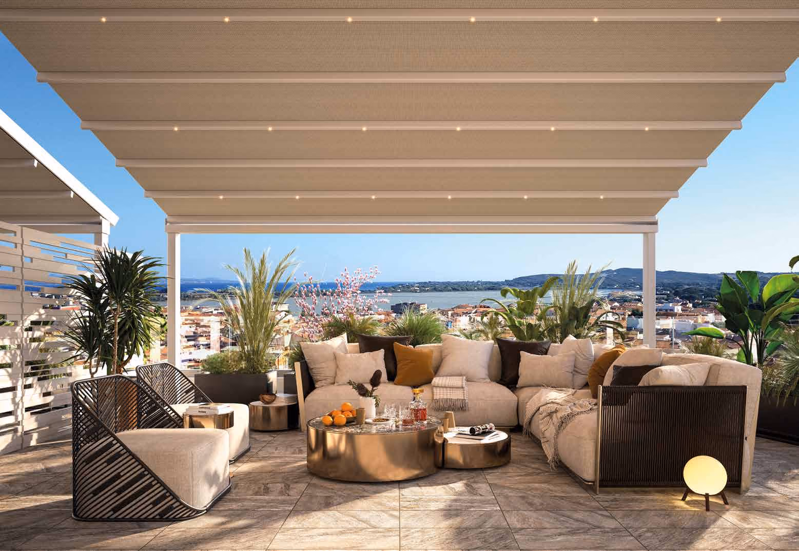 Modern outdoor lounge area under a large pergola with integrated lights, featuring beige cushioned sectional sofa with pillows, two minimalist armchairs, gold-toned round coffee tables with drinks and fruits, surrounded by potted green plants, overlooking a coastal cityscape and blue sky.