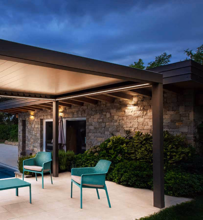 Modern outdoor patio with a metal pergola, two turquoise chairs with beige cushions, and a matching coffee table on tiled floor next to a house with stone walls and greenery, under a dusky sky.
