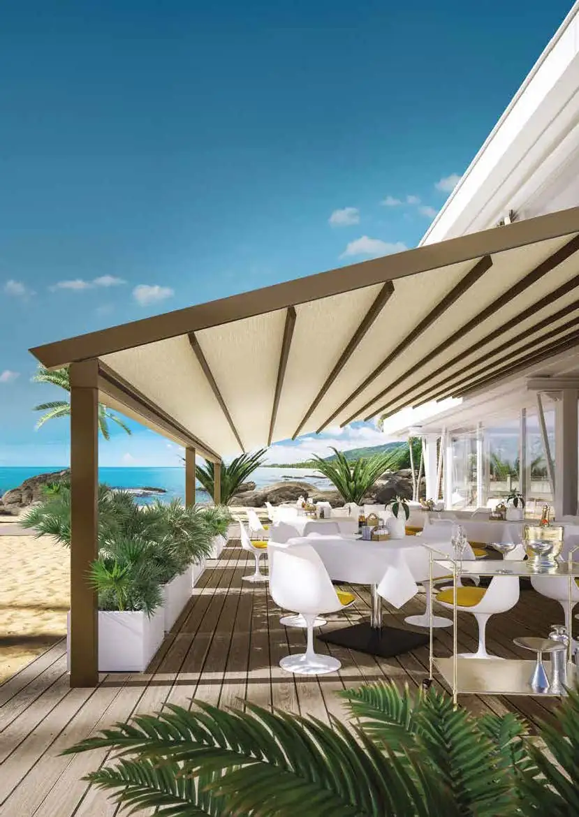 Outdoor patio dining area on wooden deck with white tables and chairs under a beige pergola, overlooking a sandy beach with palm trees and ocean in the background.