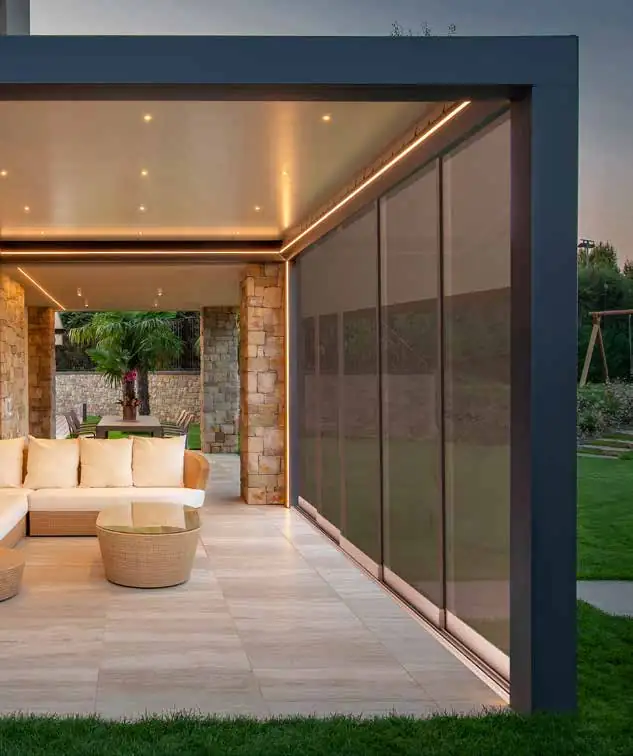 Modern outdoor covered patio with beige cushioned sectional sofa and round wicker coffee table, stone pillars, integrated ceiling lights, and retractable screen on one side, surrounded by green lawn and garden.