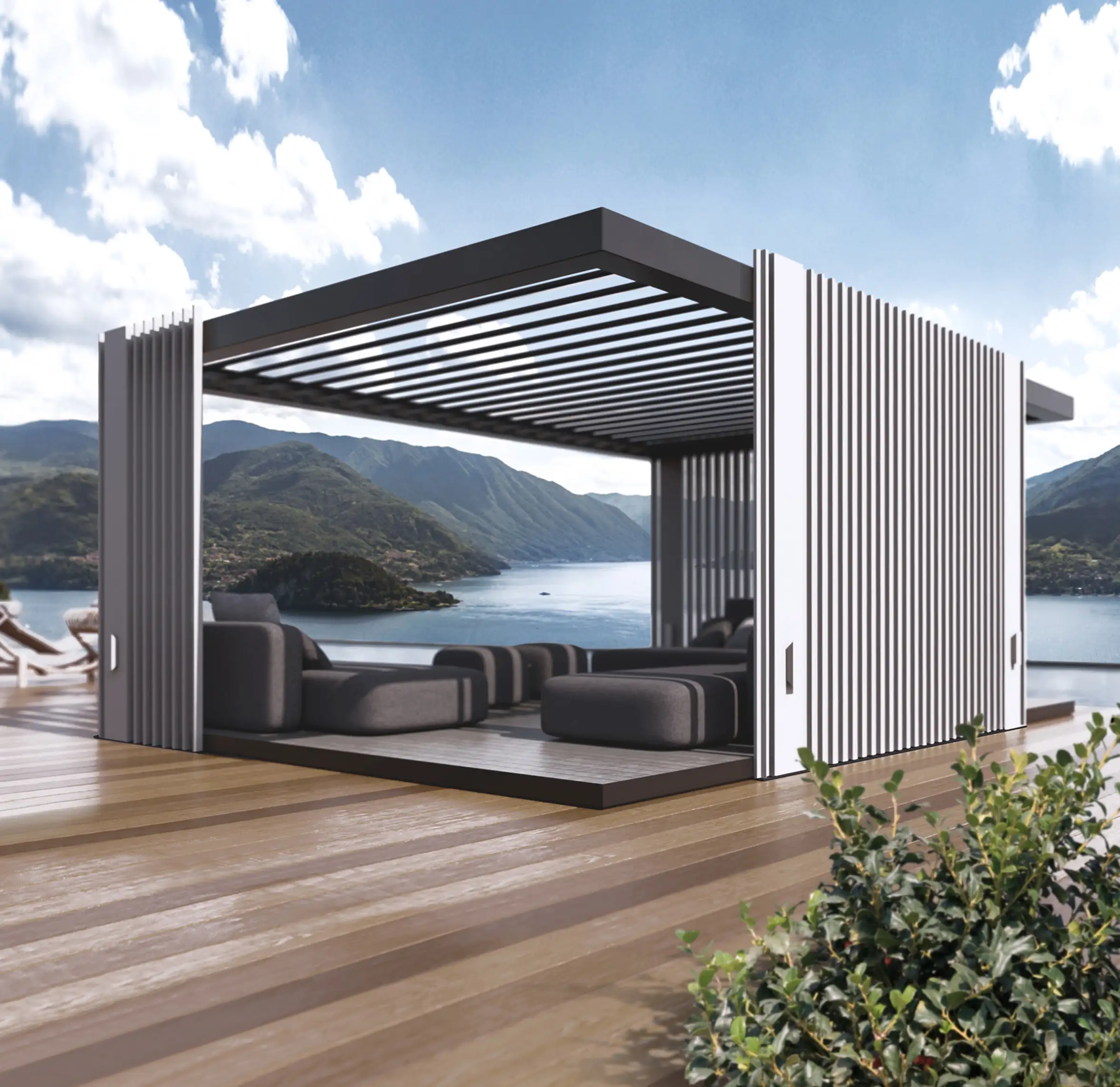 Modern outdoor lounge with grey cushioned seating under a black pergola structure with white vertical slats, overlooking a scenic lake surrounded by green mountains under a partly cloudy sky.