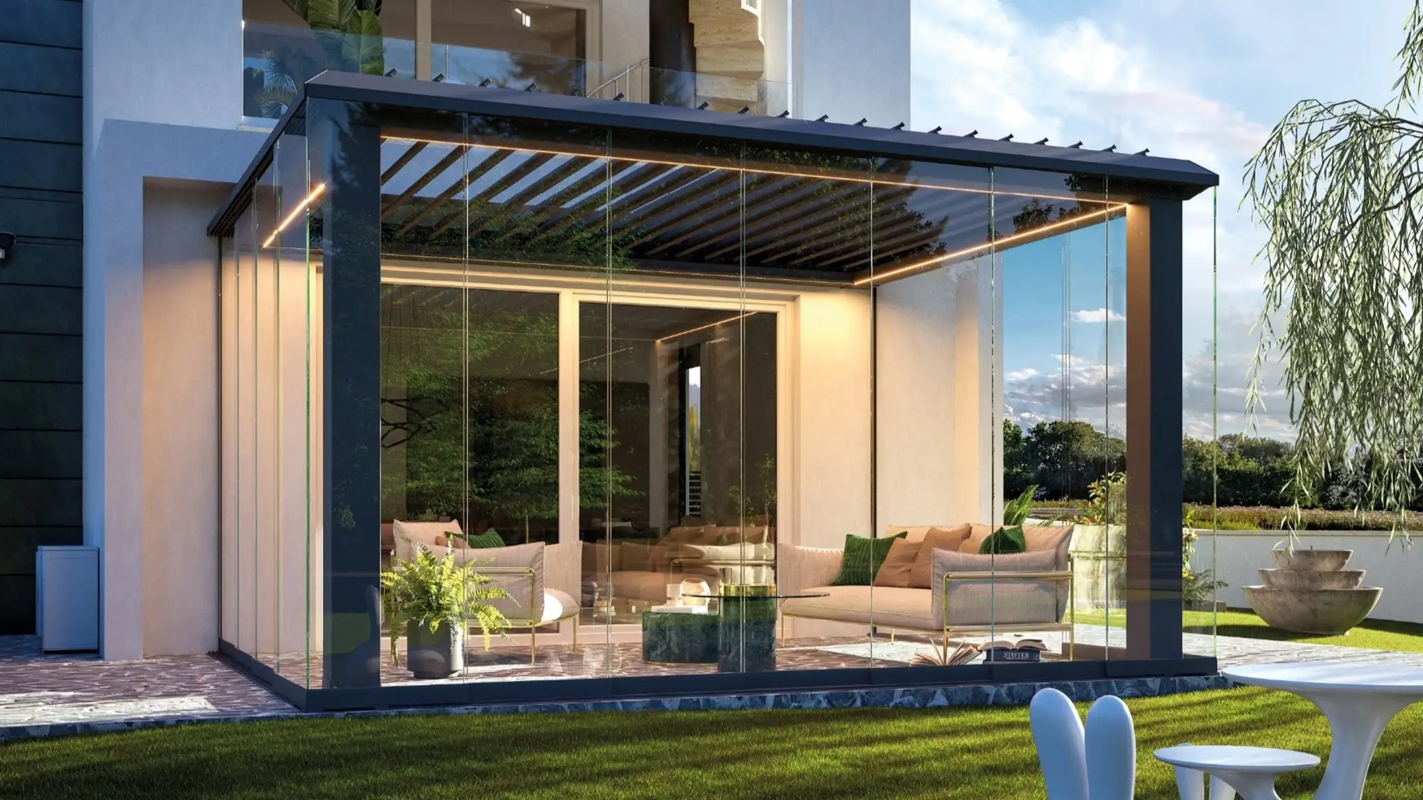 Modern outdoor glass-enclosed patio attached to a house, featuring beige cushioned sofas with green and beige pillows, a glass coffee table, and a potted fern, with a grassy lawn and outdoor furniture in the foreground under a partly cloudy sky.