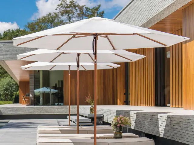 Three white patio umbrellas aligned over beige cushioned lounge chairs on a stone patio next to a modern building with wooden paneling, under a partly cloudy sky.