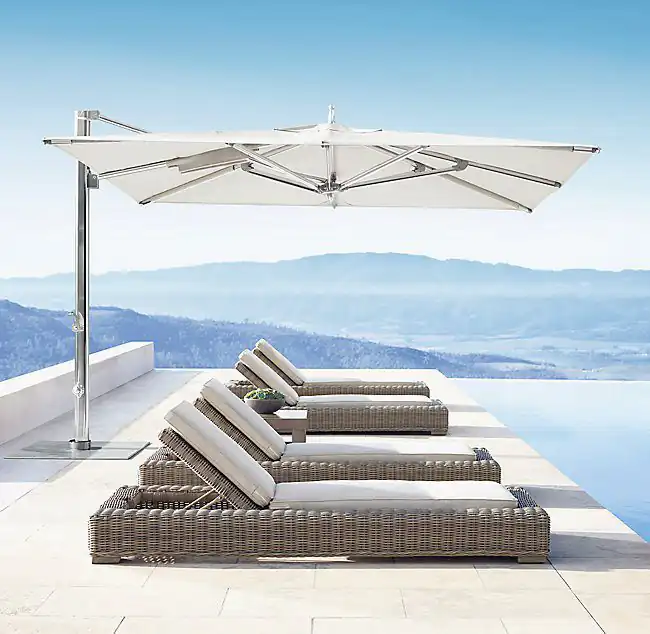 Three wicker chaise lounges with white cushions arranged in a row beside an infinity pool, shaded by a large modern cantilever umbrella with a metal pole, overlooking distant blue mountains under a clear sky.