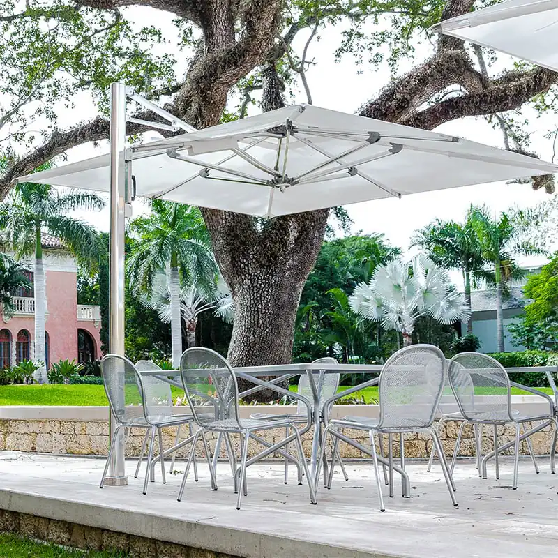 Outdoor patio area with metal mesh chairs and a glass-top table under a large, rectangular white cantilever umbrella, surrounded by trees and greenery near a stone wall and residential buildings.