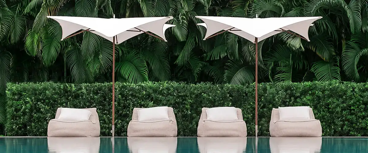 Four beige cushioned lounge chairs with white umbrellas behind a calm swimming pool, set against dense green tropical foliage.