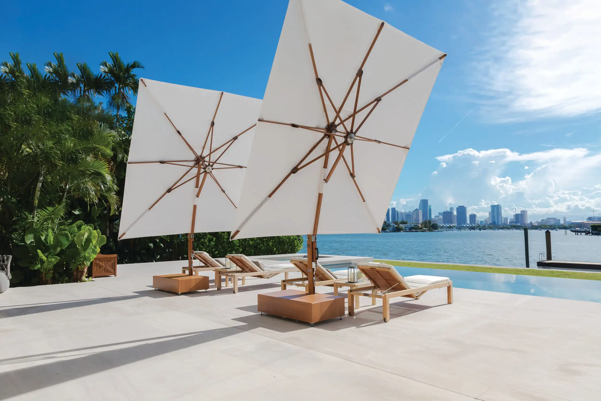 Two wooden lounge chairs with white cushions under large white umbrellas on a spacious patio by an infinity pool, overlooking a body of water and a city skyline under a blue sky.