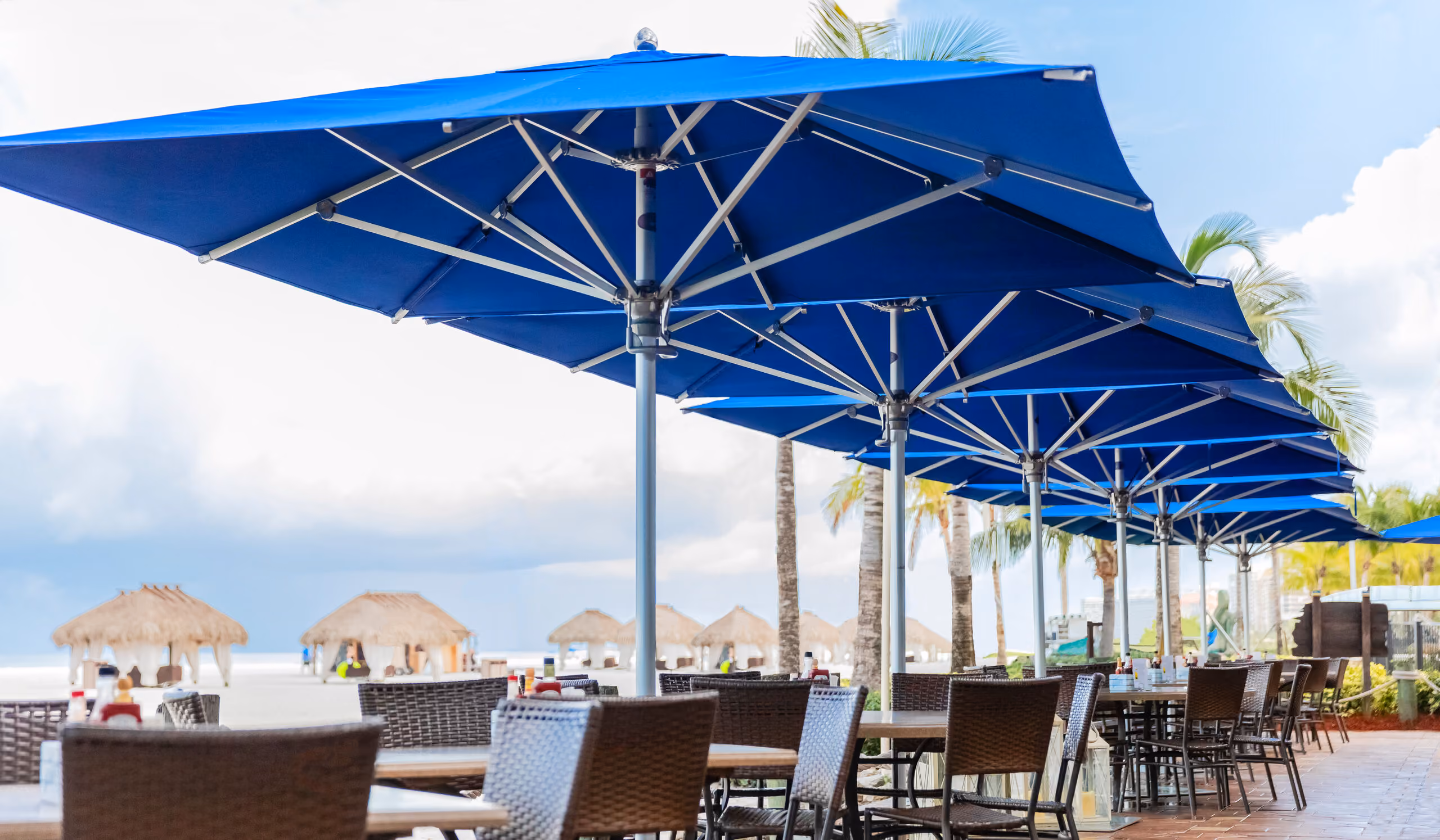 Outdoor dining area with multiple tables and chairs under large blue umbrellas near a beach with palm trees and thatched-roof huts in the background.