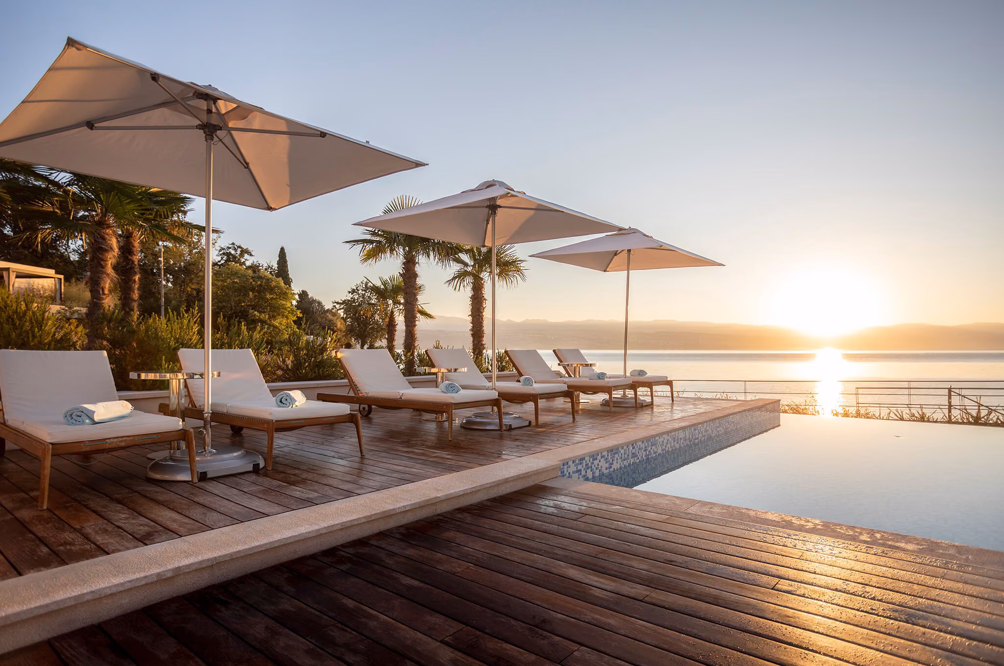 Sunset over an infinity pool with wooden deck featuring six white cushioned lounge chairs each with a rolled towel, shaded by large white umbrellas, surrounded by palm trees and greenery.