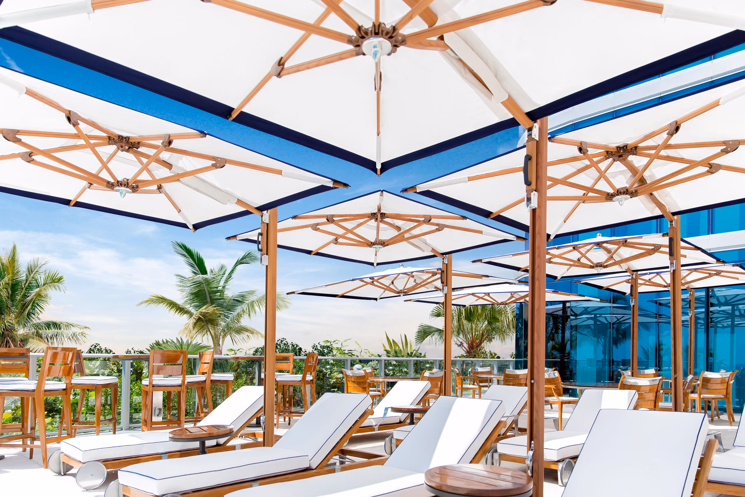 Outdoor lounge area with white cushioned sun loungers under large white umbrellas with wooden frames, surrounded by wooden tables and chairs, glass railing, and palm trees in the background under a blue sky.