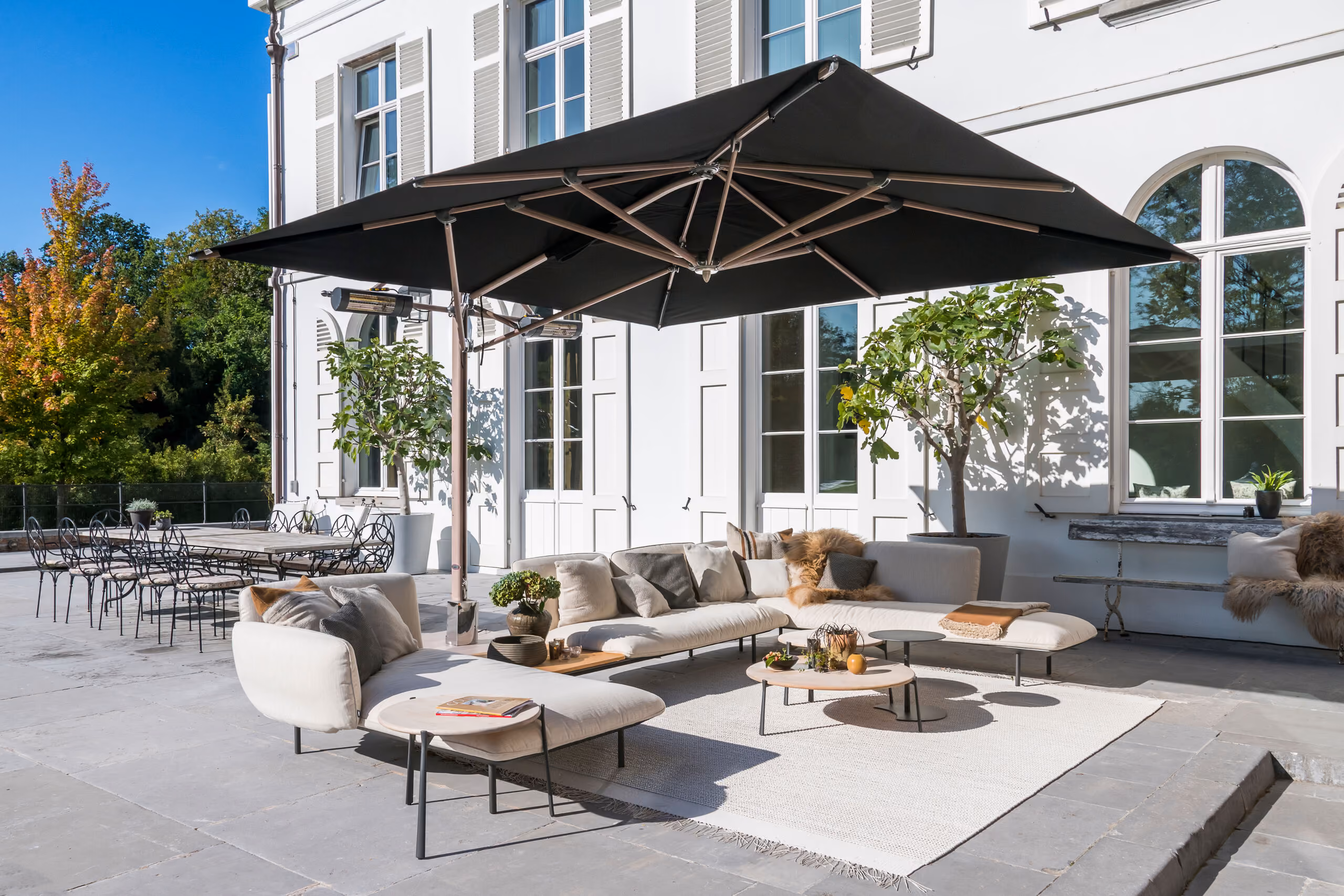 Modern outdoor patio with a large black umbrella shading a beige sectional sofa with cushions, a round coffee table, and a white rug, set against the white exterior wall of a building with tall windows and potted plants.