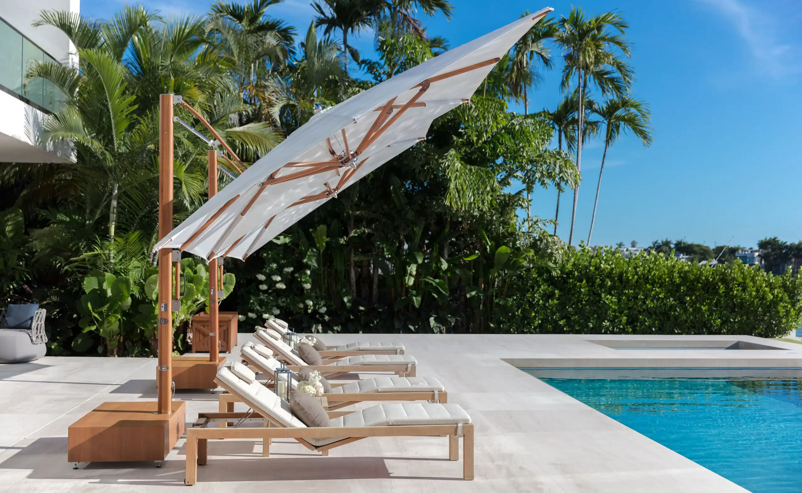 Poolside scene with five cushioned wooden lounge chairs under a large angled white canopy umbrella, surrounded by lush green palm trees and bushes, next to a clear blue swimming pool under a bright sunny sky.