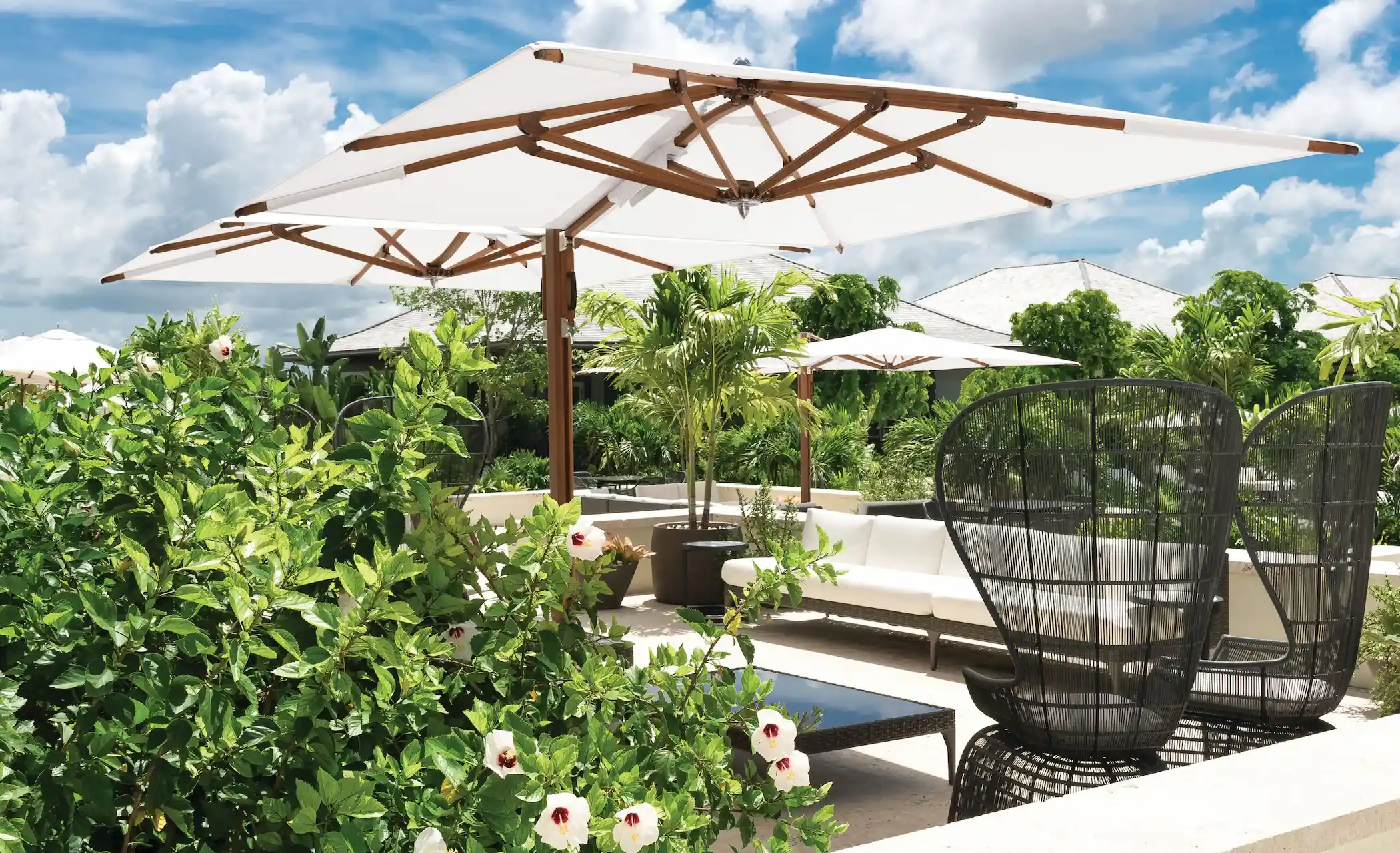 Outdoor seating area with white cushioned sofas, black woven chairs, wooden-framed white umbrellas, surrounded by lush green plants and hibiscus flowers under a partly cloudy blue sky.