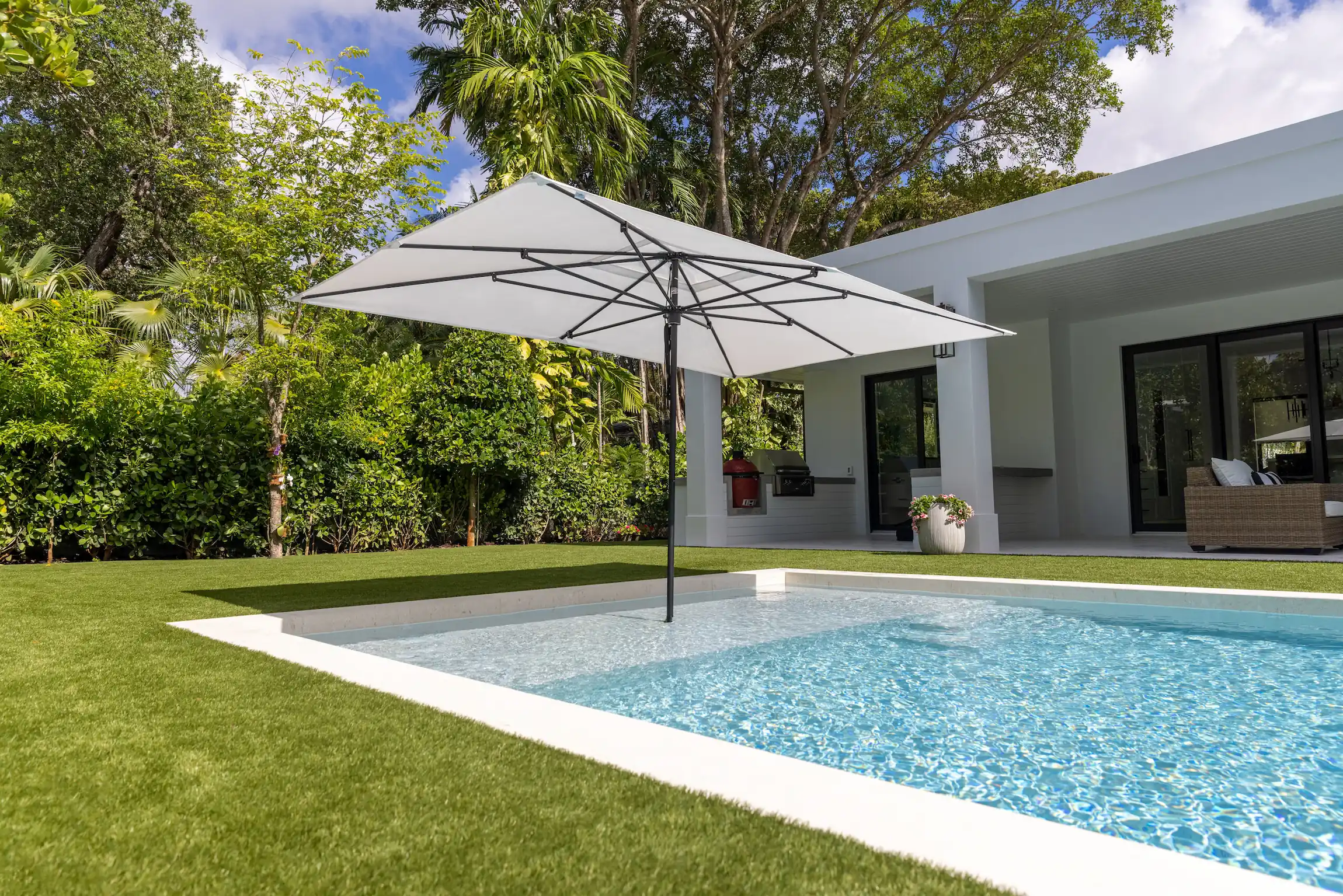 Modern backyard with a rectangular pool featuring clear blue water and a large white square umbrella anchored in the pool, surrounded by green grass, dense trees, and a white house with a shaded patio and outdoor furniture.