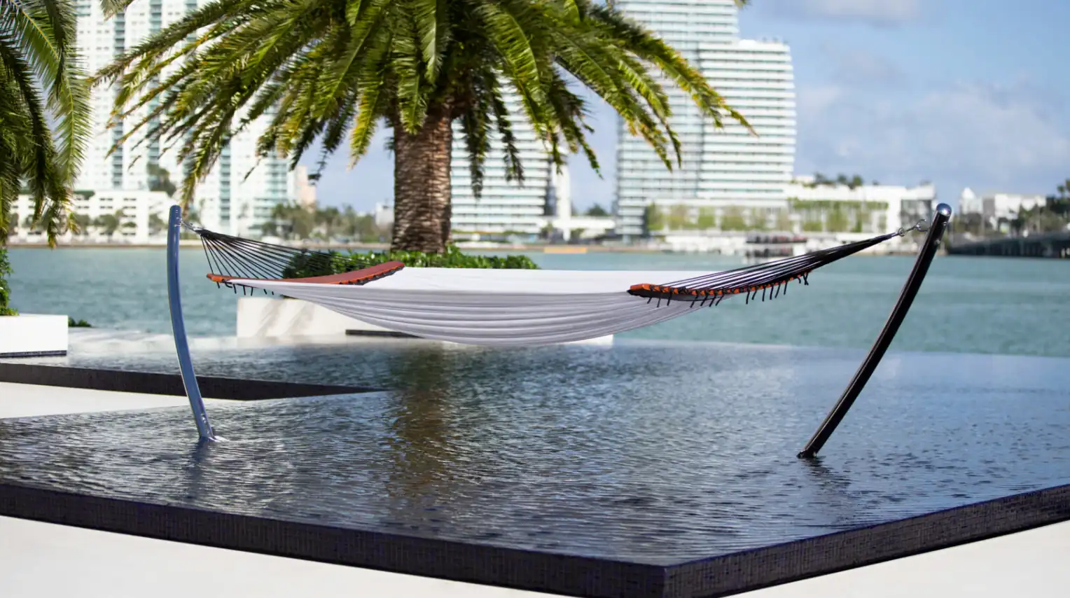 White hammock with black and orange accents suspended over a shallow water pool, with palm trees and modern high-rise buildings in the background by a waterfront.