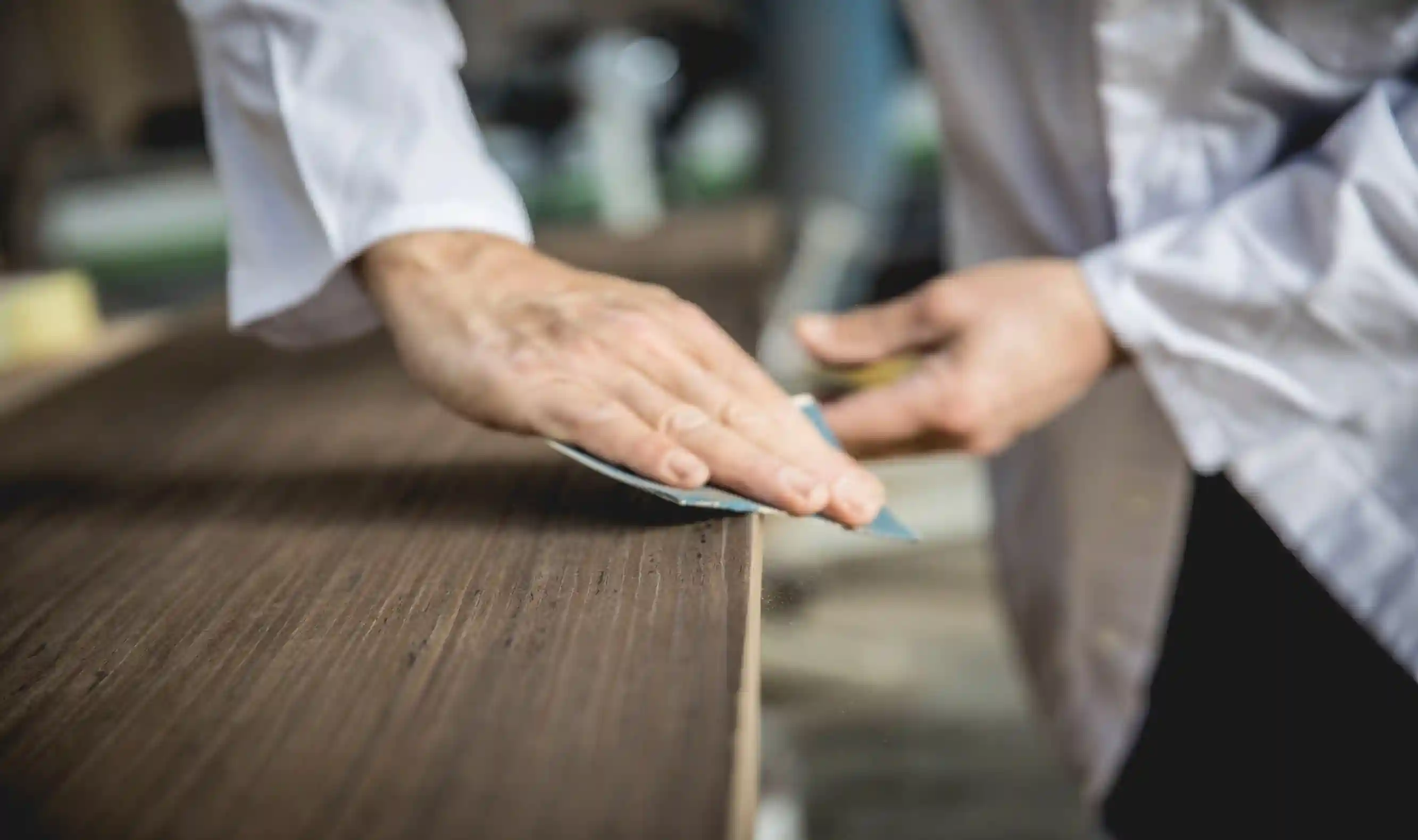 Person smoothing a wooden surface with sandpaper, focusing on the hand and wood edge.