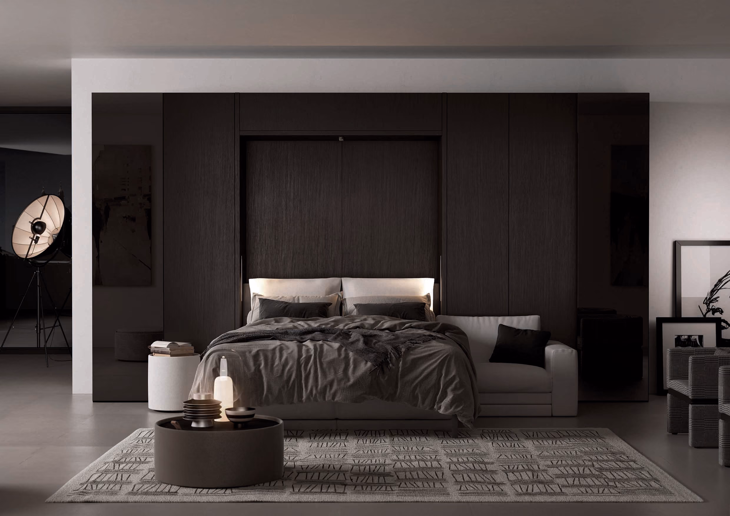 Modern bedroom with a large dark wood headboard, a double bed with gray bedding and pillows, a white couch on the right, a round black coffee table with decorative items, and a patterned rug on the floor.