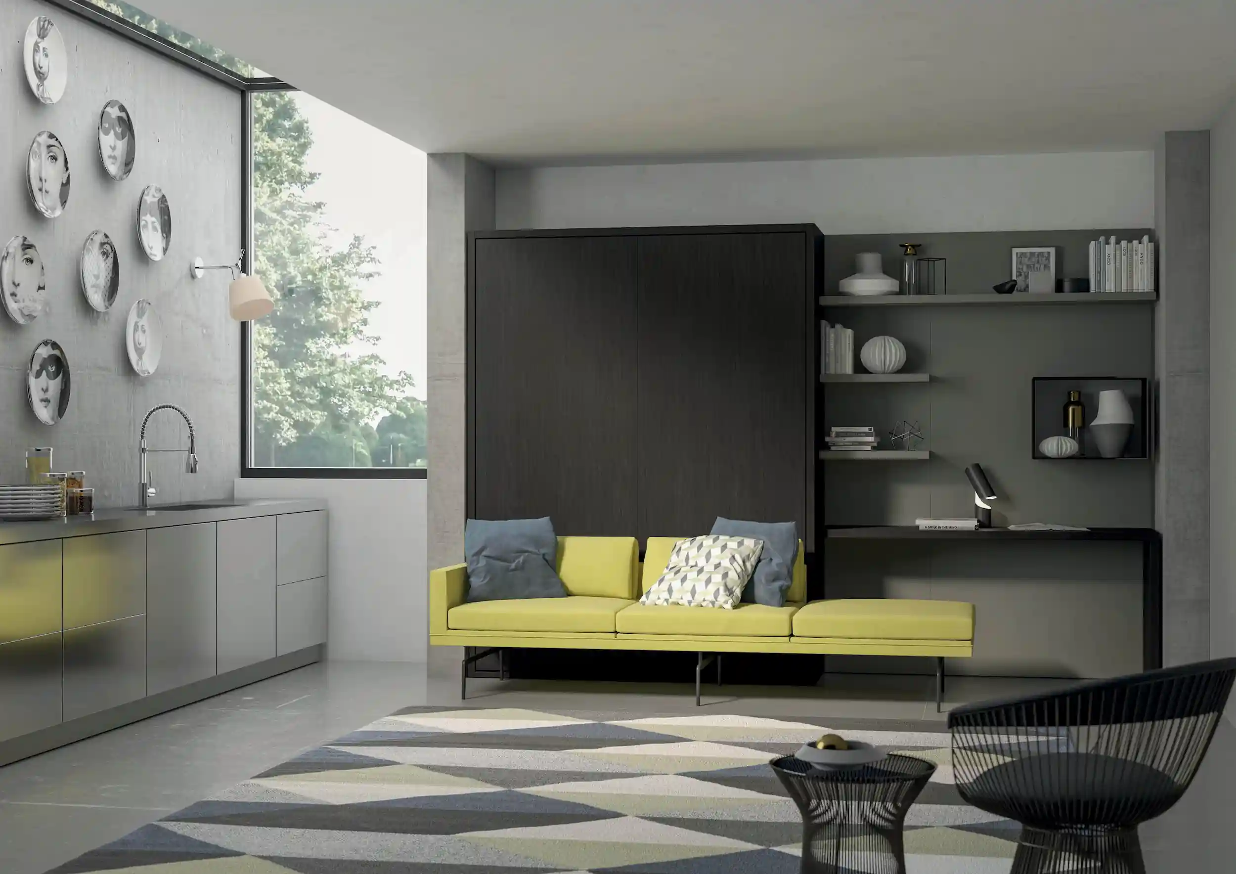 Modern living space with a yellow sofa adorned with blue and patterned pillows in front of a dark wood cabinet, adjacent to a kitchen area with gray cabinets and a wall decorated with artistic face plates. A geometric patterned rug covers the floor, and black wireframe chairs and a small table are in the foreground. Shelves with books and decorative objects flank a built-in desk with a small lamp on the right wall. A large window offers a view of green trees outside.