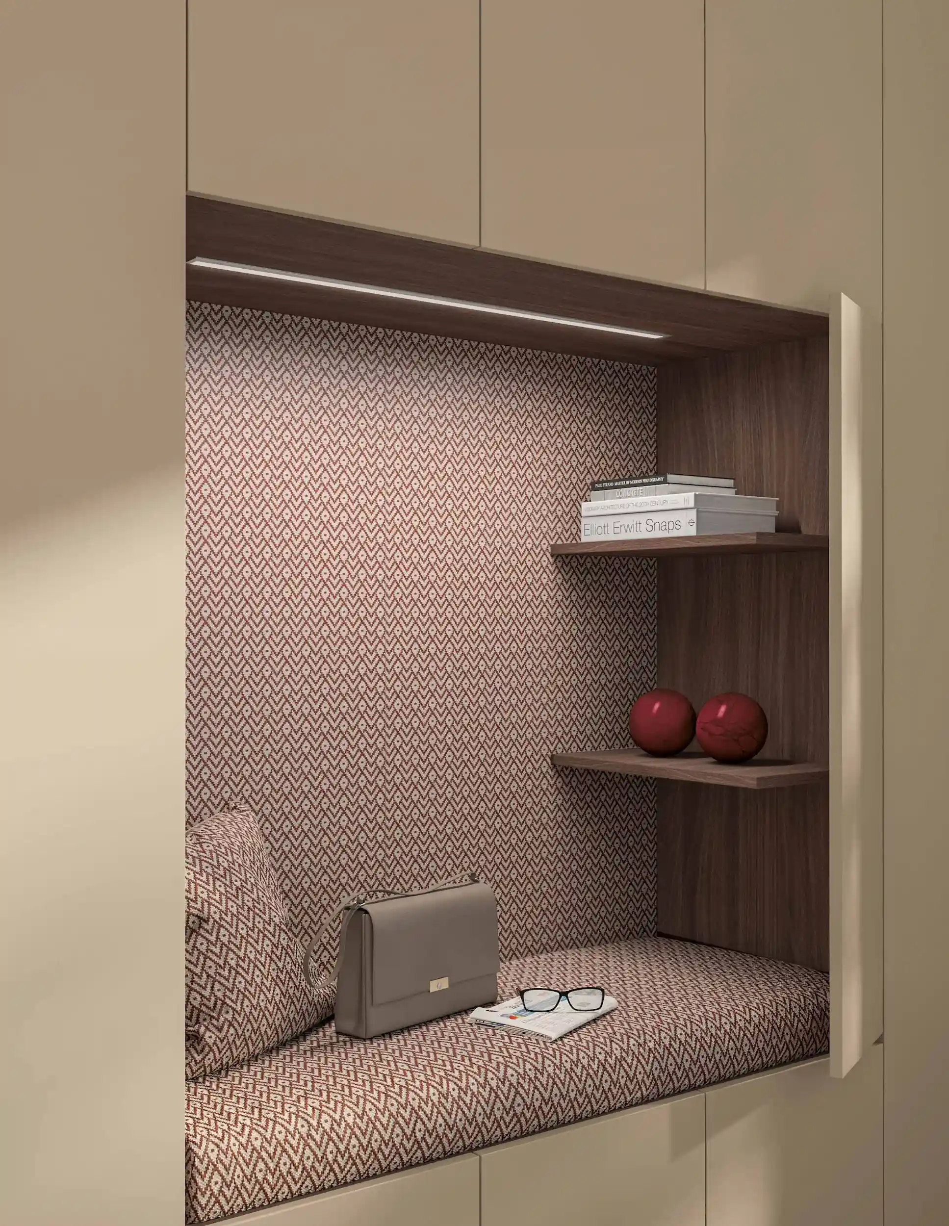 Built-in beige nook with a patterned cushioned seat and backrest, a gray handbag, eyeglasses on an open book, wooden shelves holding stacked books and two red decorative spheres, and overhead LED lighting.