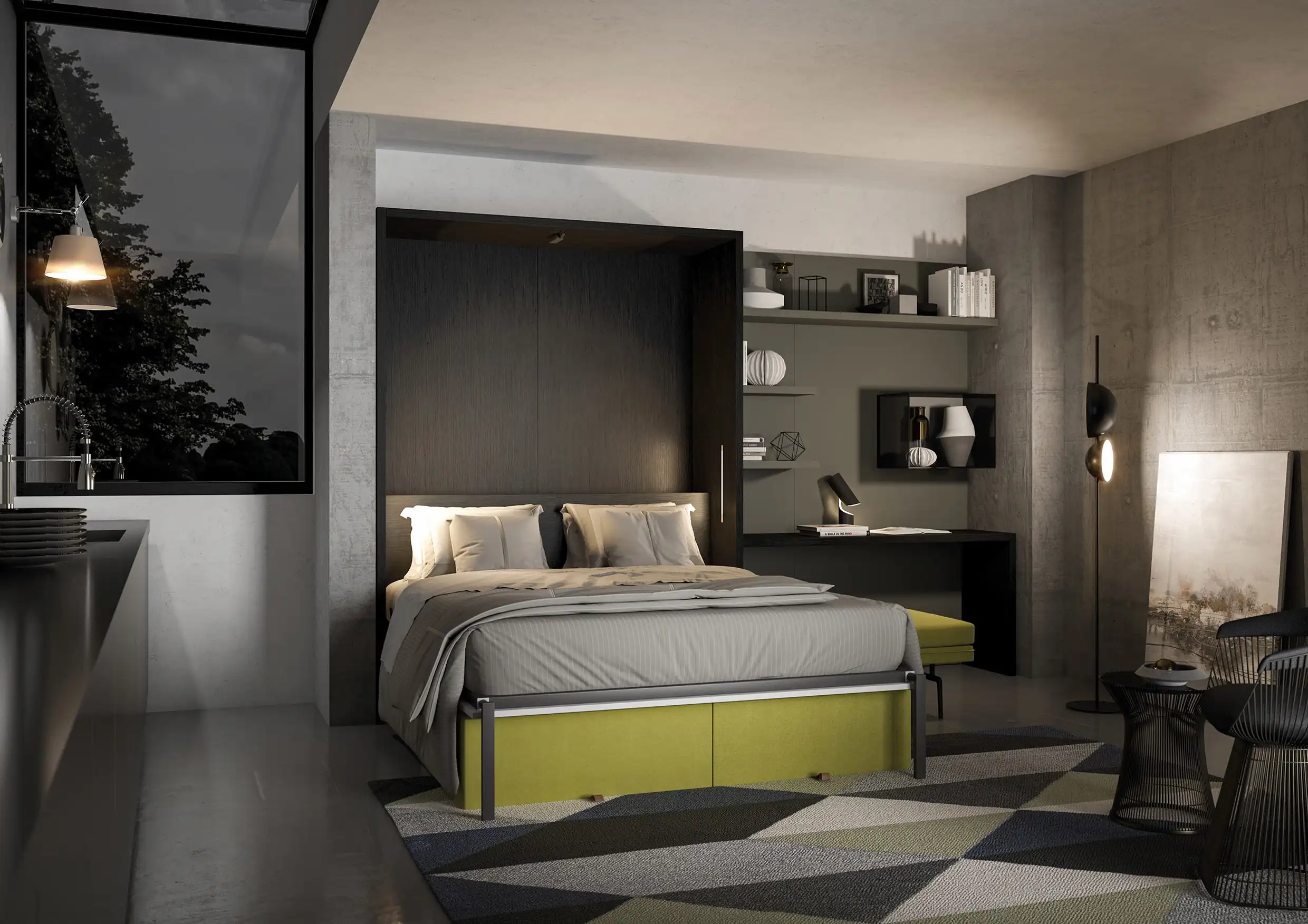 Modern bedroom with a large bed featuring gray bedding and white pillows, set in a dark wood recessed frame; a patterned rug with geometric shapes in gray, green, and beige covers the floor. To the right, there is a black desk with shelves holding books and decorative items, a small bench with green upholstery, a floor lamp with two lights, and a piece of artwork leaning against a concrete wall. To the left, a window shows dark trees outside and a hanging lamp next to a sink area.