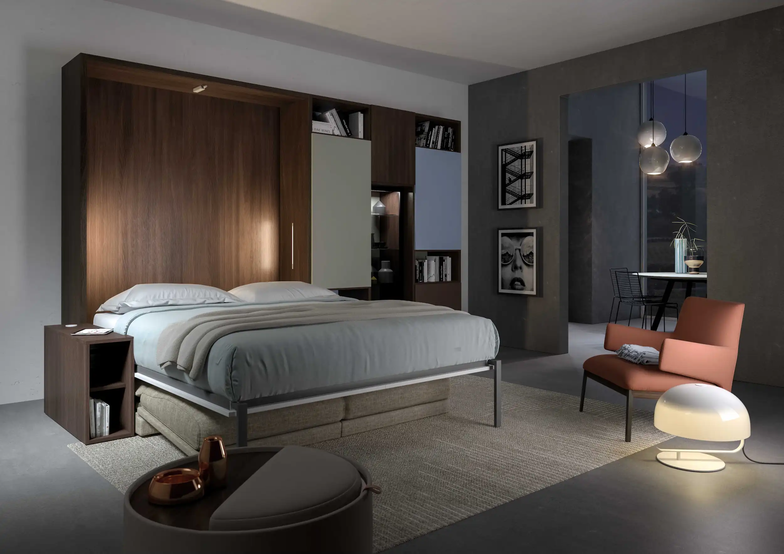 Modern bedroom with a large bed featuring gray bedding and a wooden headboard with built-in shelving and cabinets, a small side table, a coral armchair with a folded blanket, a white modern floor lamp, and framed black-and-white art on a charcoal gray wall. A dining area with hanging pendant lights and a small table with chairs is visible through an open doorway.