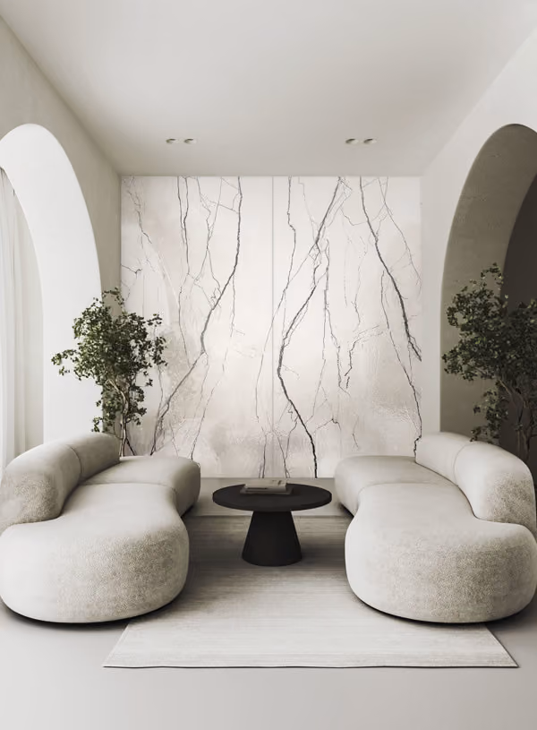 Minimalist living room with two curved cream sofas facing each other around a low black round coffee table, set on a light rug, with large marble slabs with dark veins on the back wall and two green plants in the corners under arched openings.