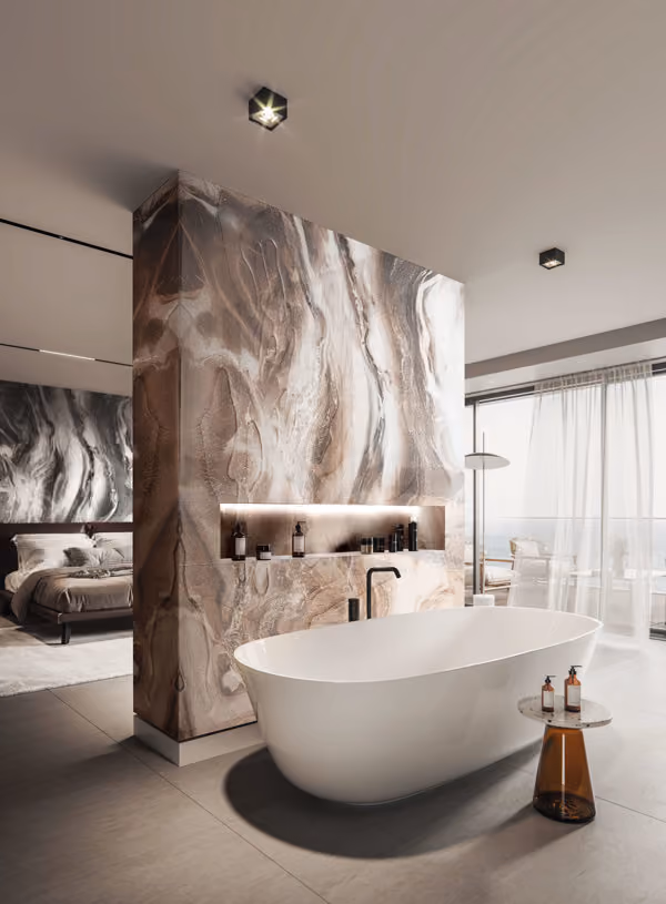 Modern bathroom with a free-standing white bathtub in front of a large marble-patterned wall that separates the bathroom from a bedroom. The wall features a recessed shelf with bottles and LED lighting. A small round side table next to the bathtub holds two bottles. The space includes large floor-to-ceiling windows with sheer curtains, allowing natural light and a view outside.