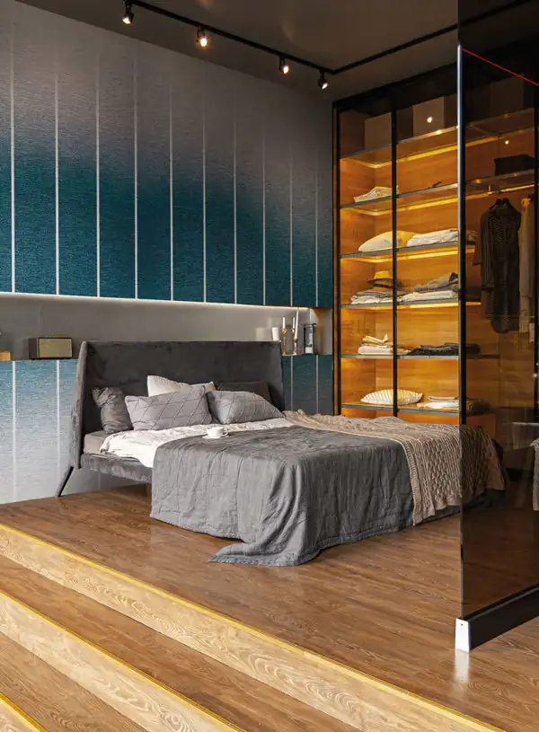 Modern bedroom with a gray upholstered bed on a raised wooden platform with steps, featuring gray bedding and pillows, a textured gradient blue wall behind, and a glass-enclosed wooden wardrobe with folded clothes lit by warm lighting.