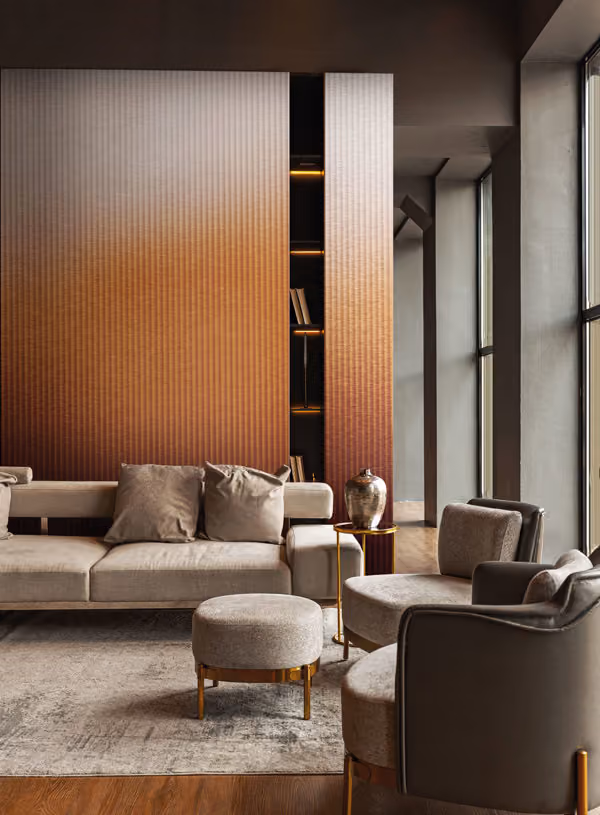 Modern living room with beige sofa and matching armchairs around a round ottoman, featuring a large textured wooden wall panel, tall windows, and a metallic vase on a small side table.