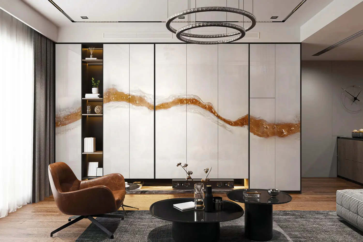 Modern living room featuring a brown leather armchair on a gray rug, two black round coffee tables with decorative items, and a large wall unit with a flowing bronze-colored abstract design across the white panels, next to built-in shelves with books and decor, under a circular double-ring chandelier.